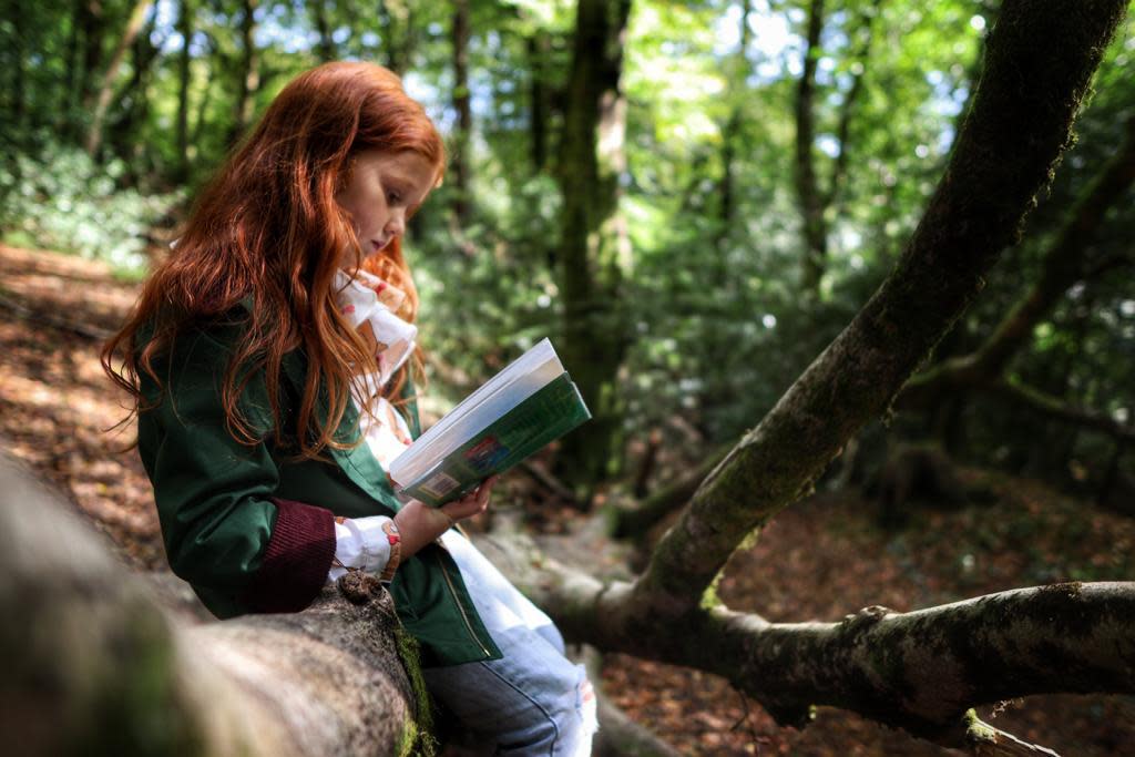 A girl with red hair reading in a woods.