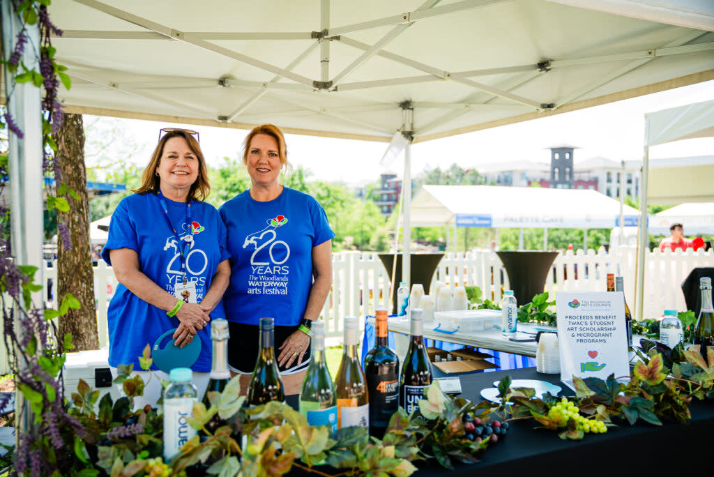 Two women volunteers at the WWAF (wearing matching, bright blue, "20 Years" shirts for the festival) smile for the camera as they work at a wine tasting booth.
