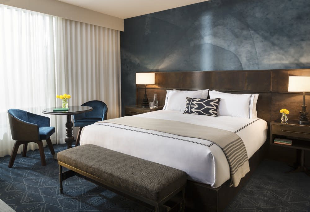 Room in the Hotel Van Zandt. The bed is covered in white linens and a beige blanket, there is a wooden headboard and blue textured paint behind the wall and a small table with two chairs at the bedside