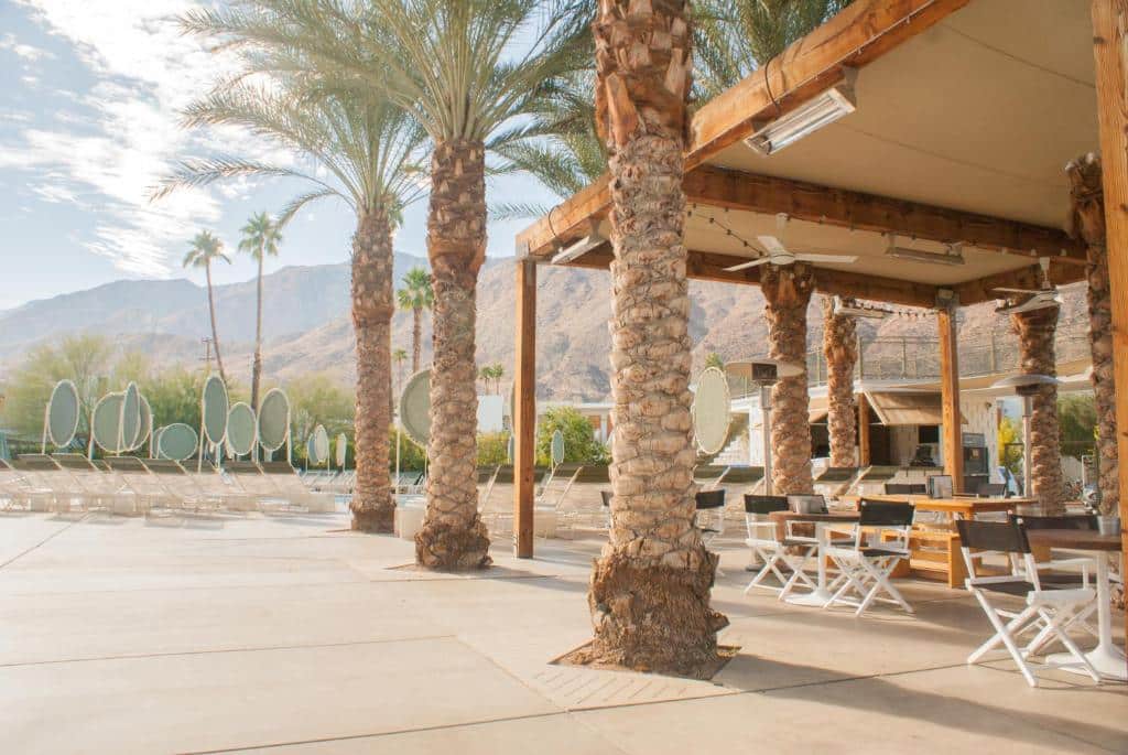A sunny poolside area with lounge chairs, palm trees, and a mountainous backdrop, adjacent to a shaded pavilion with seating.