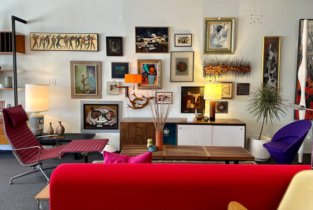 An eclectic art gallery interior with various framed paintings adorning the walls, modernist-style furniture including a red chaise longue and a purple chair, floor lamps, decorative vases, and a tall plant pot.
