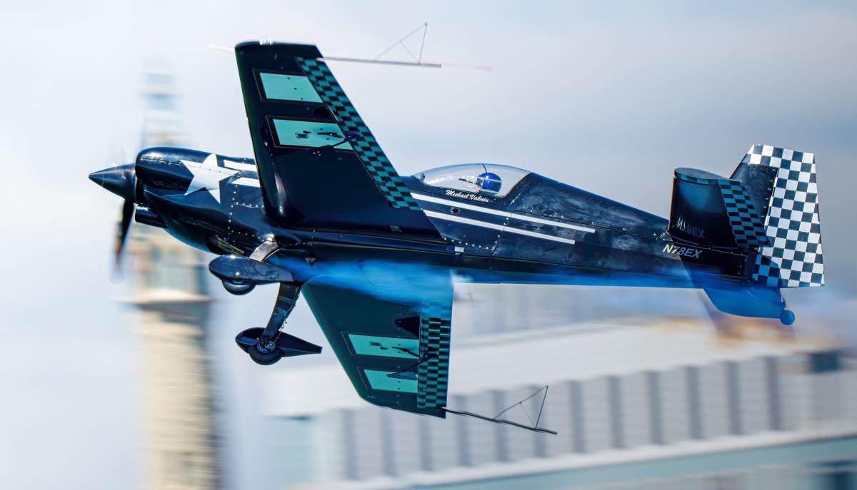A blue aerobatic airplane performs a low-altitude maneuver over water, creating a mirrored reflection on the calm surface below. Motion blur in the background emphasizes the aircraft’s speed and precision during an air show performance.