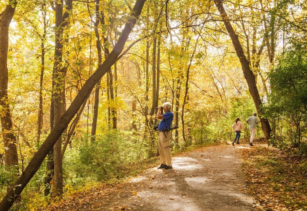 man is looking through binoculars at the park. Two people playfully run along the track away from the camera.