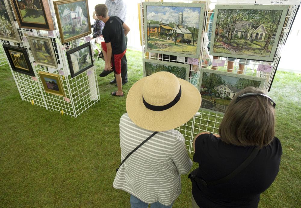 Woman in tan hat and white sweater and woman in black sweater look at completed art paintings part of Paint Dexter Plein Air.