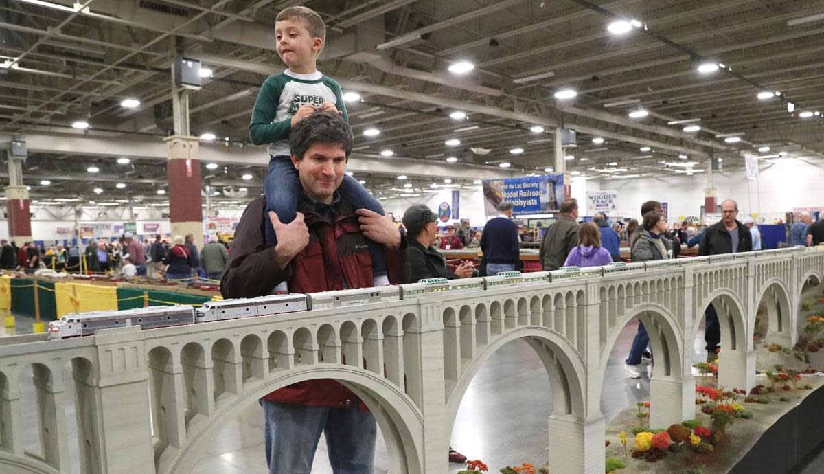 A man carries a young boy on his shoulders as they admire a large model train display at Trainfest. The model train runs across a detailed arched bridge, while other visitors walk and observe exhibits in the busy convention hall.