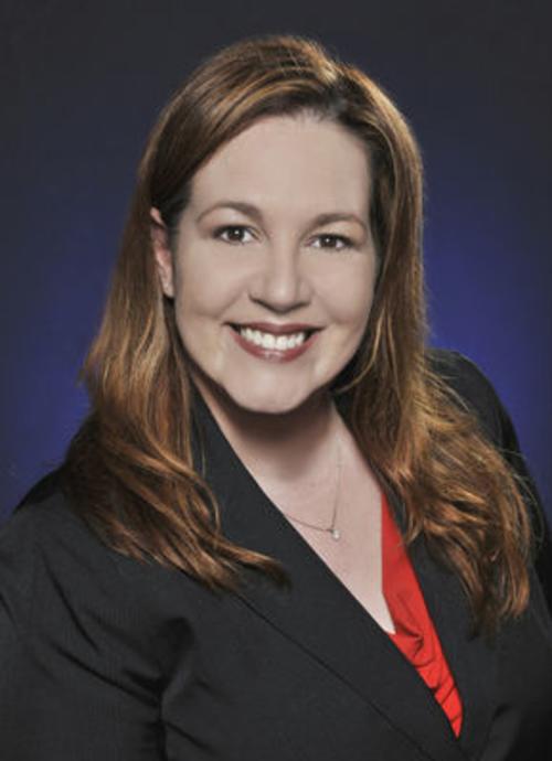 Professional headshot of a woman with long auburn hair wearing a black blazer over a red top, smiling against a dark gradient background.