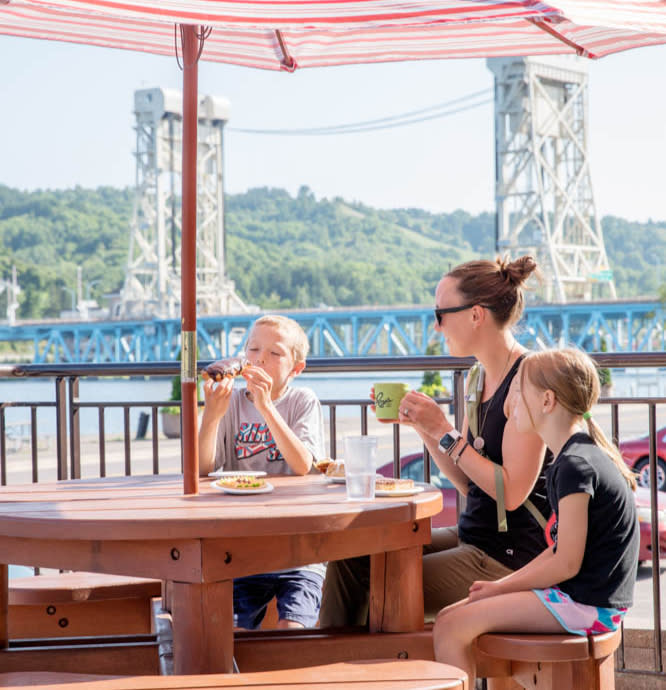 Two children and adult eat at outside picnic table with Portage Lake Lift Bridge in background.