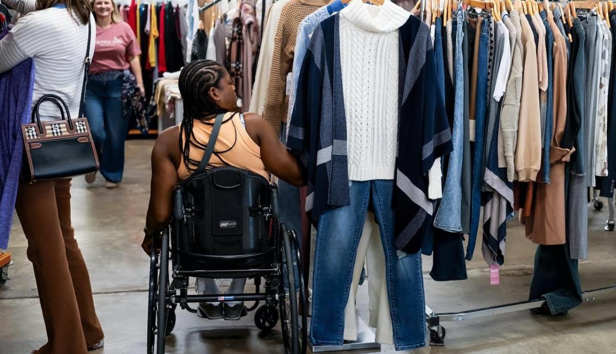 A person using a wheelchair browses clothing racks inside Flawless Consignment Boutique. They are looking at a display featuring a white cable-knit sweater layered under a navy and gray poncho, paired with blue jeans. Other shoppers and racks of assorted clothing are visible in the background.