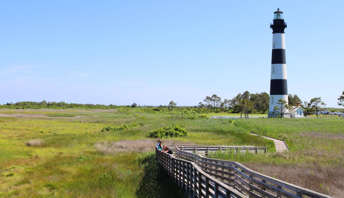 A view from the wooden walkway looking toward Bodie Island Lighthouse, surrounded by marsh on a bright sunny day.