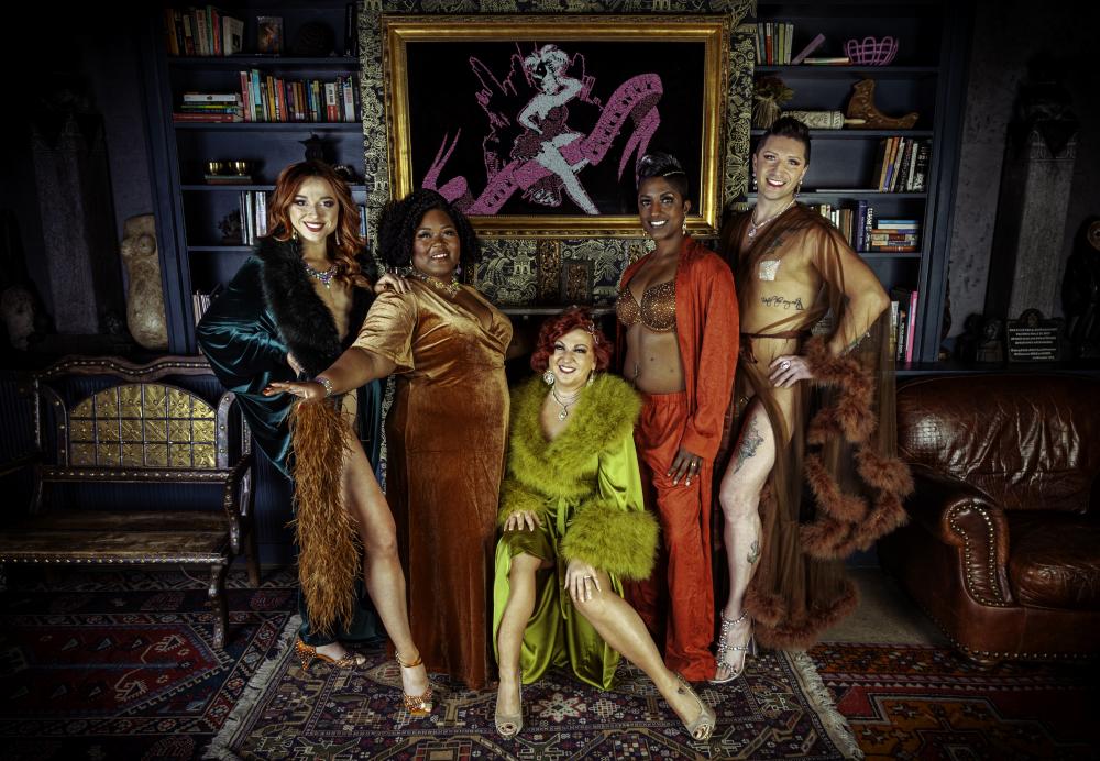 A group of 5 people all looking at a camera and smiling. They are wearing velvets, satins, and fur/feather trims. The picture implies opulence.