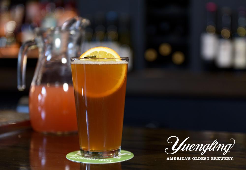 Pint glass filled with beer in front of a pitcher of blood orange juice sitting on top of a brown bartop