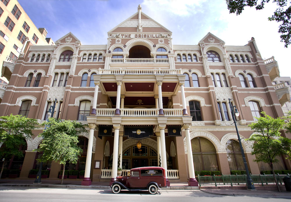 Four-story exterior of the Sixth Street Entrance at the historic Driskill Hotel. There is a red antique car parked in front of the columned entrance