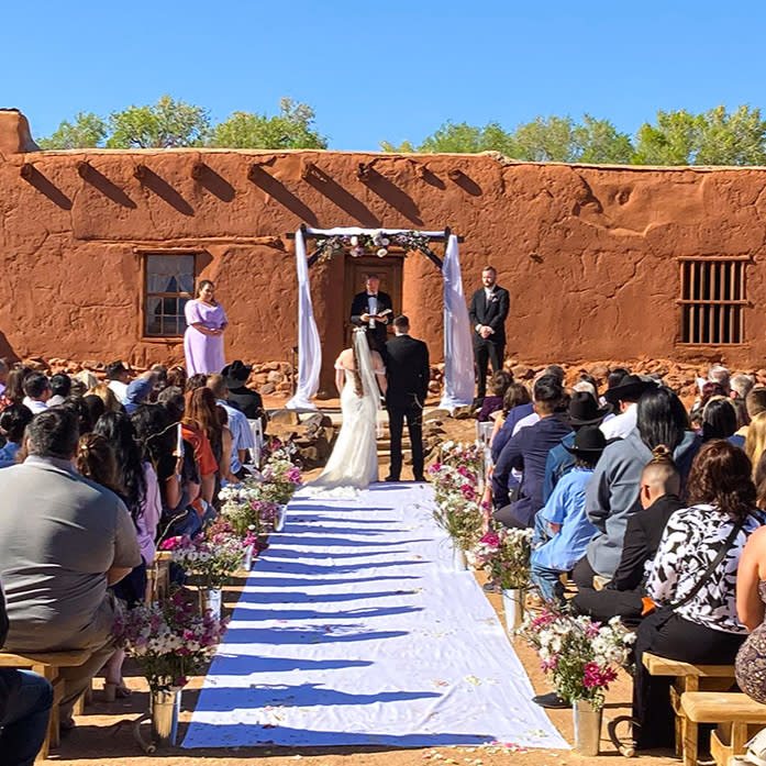 Wedding ceremony outdoors with a couple under an arch, guests seated on wooden benches. The backdrop is an adobe building, clear blue sky.