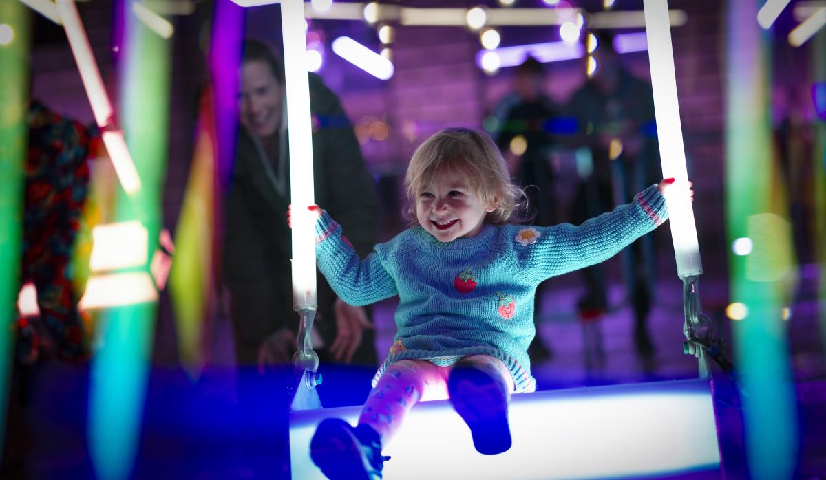 A little girl enjoys playing on an illuminated swing, with neon lights in the background