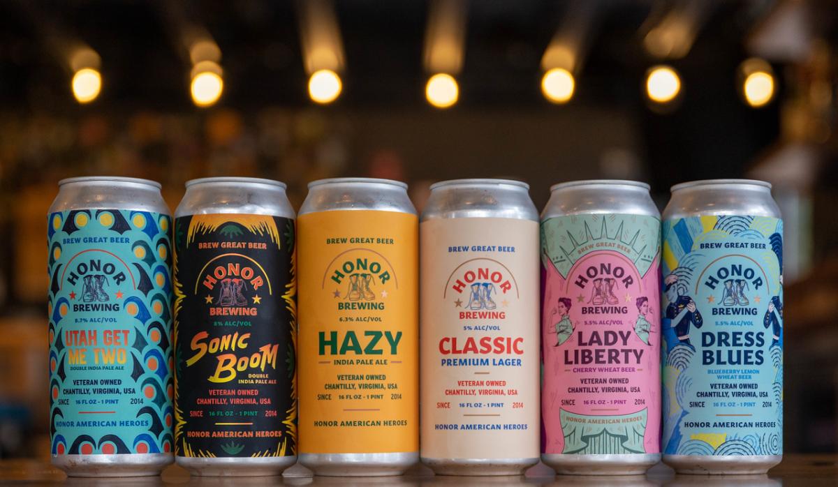 Six colorful beer cans from Honor Brewing are displayed on a wooden surface, with warm lights in the background.