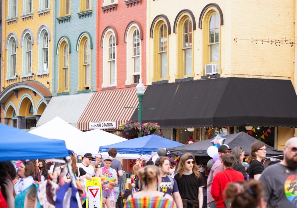 Several people walk around during a street festival with vendor booths lining the streets. Behind vendor booths are colorful buildings lined in a row: yellow, teal, red, and yellow