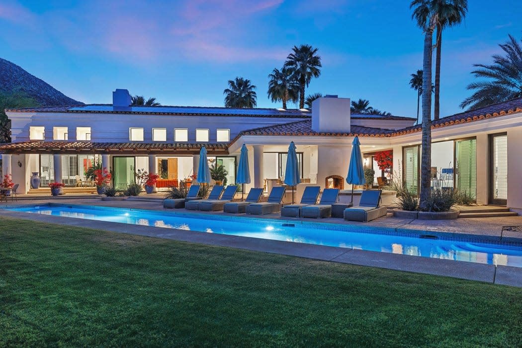 A luxurious single-story house with terracotta roofing and a row of sun loungers by a swimming pool during twilight with nearby palm trees and mountains in the background.