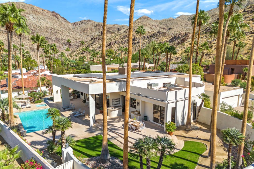 Aerial view of a modern luxury house with a swimming pool, surrounded by tall palm trees against a backdrop of mountainous terrain under a clear blue sky.