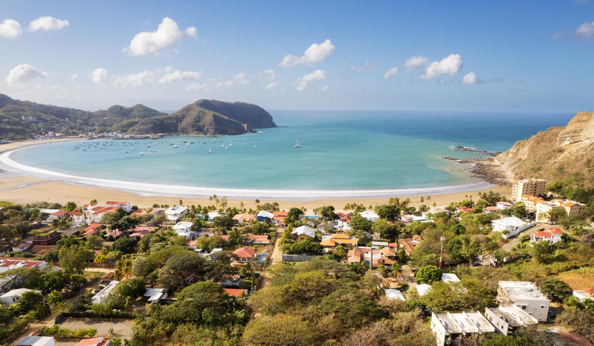 Aerial view of San Juan del Sur, Nicaragua. A crescent-shaped beach with white sand and turquoise water is surrounded by lush green hills. Boats are anchored in the bay, and colorful houses dot the landscape.