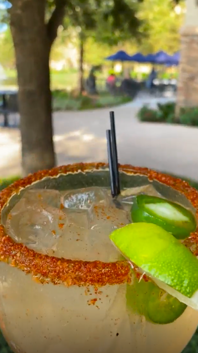 A pale yellow happy hour drink takes up the bottom half of the frame. There are sliced jalapenos floating in the drink with Tajin and a lime wedge on the rim of the glass. Behind the drink is a patio with tables shaded by blue umbrellas.