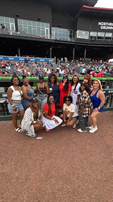 A group of attendees poses for a photo along the field-level railing at Toyota Field before a Rocket City Trash Pandas game. Fans fill the stadium seats in the background beneath the Northrop Grumman club level on a summer evening.