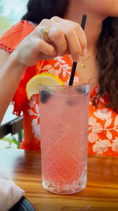 A young woman stirs a light pink drink in a tall, wide glass. There are blueberies floating in the drink and a lemon wedge on the rim. The woman is wearing a class ring and a bright orange dress with white floral embroidery.
