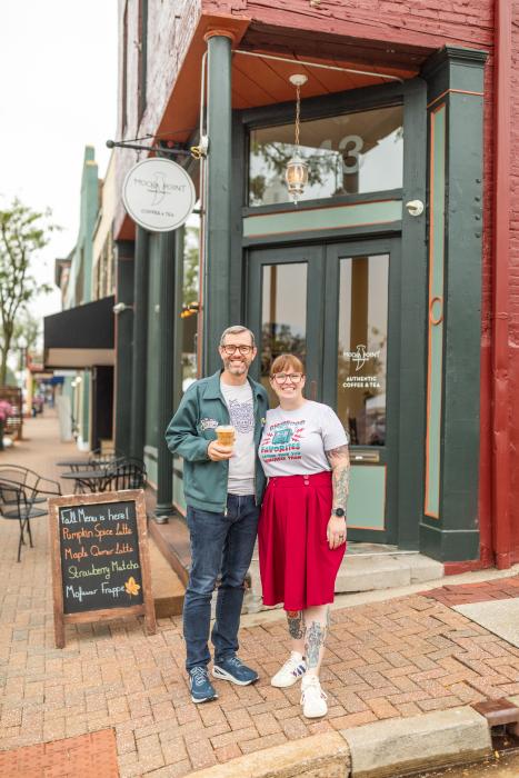 Couple standing in front of corner coffee shop in historic building that is red brick with green woodwork, man is holding an iced coffee