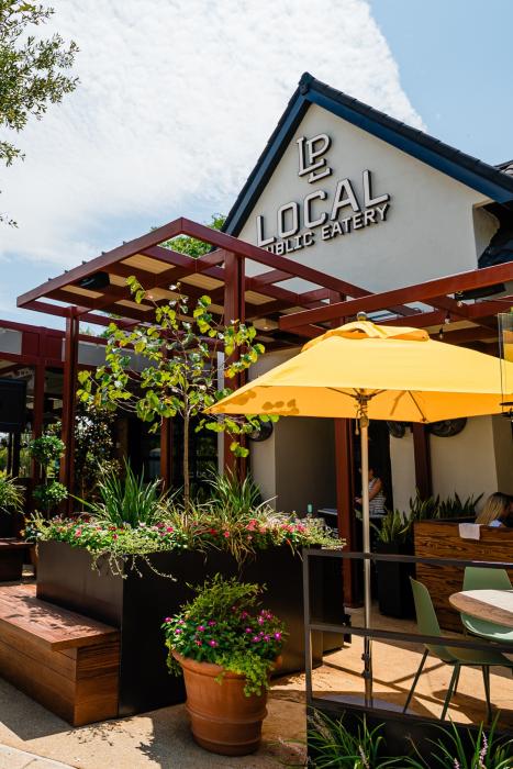 The exterior of LOCAL Public Eatery. The building features a white gable with blue trim and the name of the restaurant in large white text. The roof is gray tile, the patio awning a bright maroon metal. The patio umbrellas are a sunny yellow, the chairs a pale sage green.
