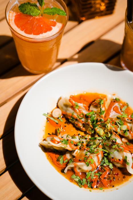 A shot of the food at LOCAL Public Eatery. On the top left is a clear plastic cup of a peachy-colored drink with a grapefruit slice and mint leaf floating on top. In the bottom right is a shallow white bowl with dumplings set like a star, drizzled in orange sauce and garnished with cilantro and green onions. The setting is on a picnic table on the patio.