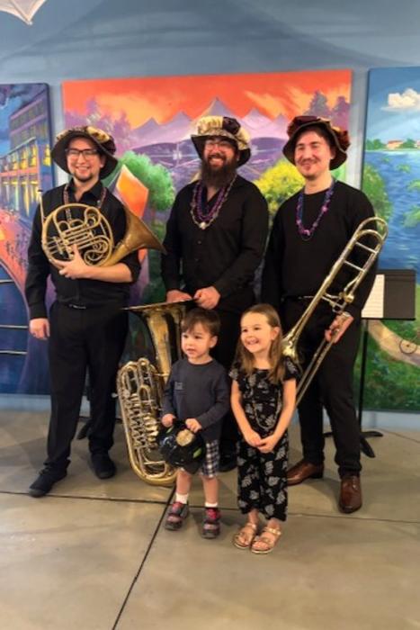 A brass trio from The Woodlands Symphony and two young children smile for the camera at The Woodlands Children's Museum. The trio is made up of three men in dark attire and Renaissance caps, each holding either a French Horn, tuba, or trombone. One of the children is a boy, the other a girl, both around 2-4 years old.