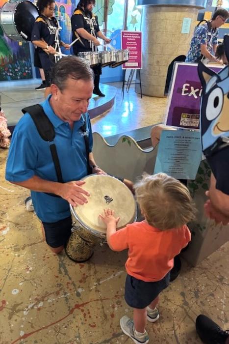 A drummer from The Woodlands Symphony kneels for a child to slap his instrument at The Woodlands Children's Museum. The man is wearing a blue shirt with the drum attached to a shoulder harness. The child is blond, wearing an orange T-shirt, dark shorts, and sneakers.