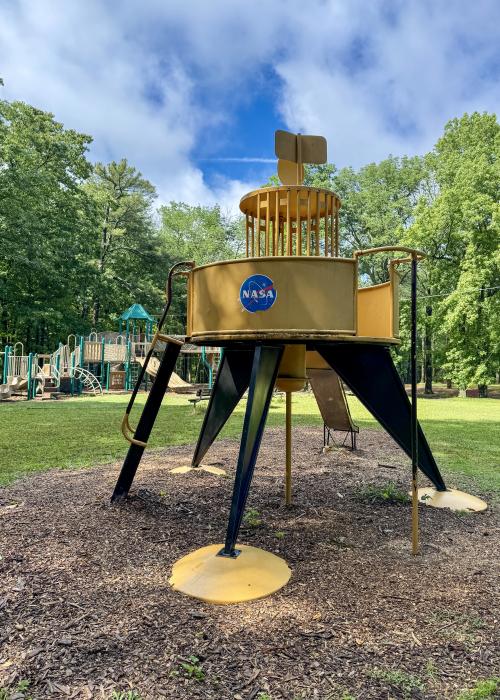 A playground structure designed to resemble a NASA lunar lander stands among the trees at Monte Sano State Park. The yellow and black play feature includes ladders and a slide, with a traditional playground visible in the background on a sunny day.