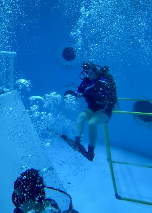 A scuba diver trains underwater in the Neutral Buoyancy Simulator at the U.S. Space & Rocket Center. Surrounded by bubbles, the diver sits on a submerged platform inside the large training tank, demonstrating an experience designed to simulate weightlessness for space-related activities.