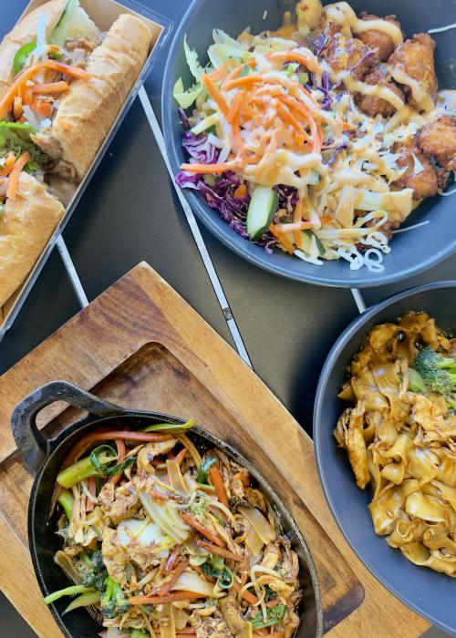 An overhead view of a variety of dishes from Viet Crispwich in Huntsville, Alabama, including a bánh mì sandwich, a crispy chicken rice bowl, stir-fried noodles, and a sizzling skillet of vegetables and noodles served on a black tabletop.
