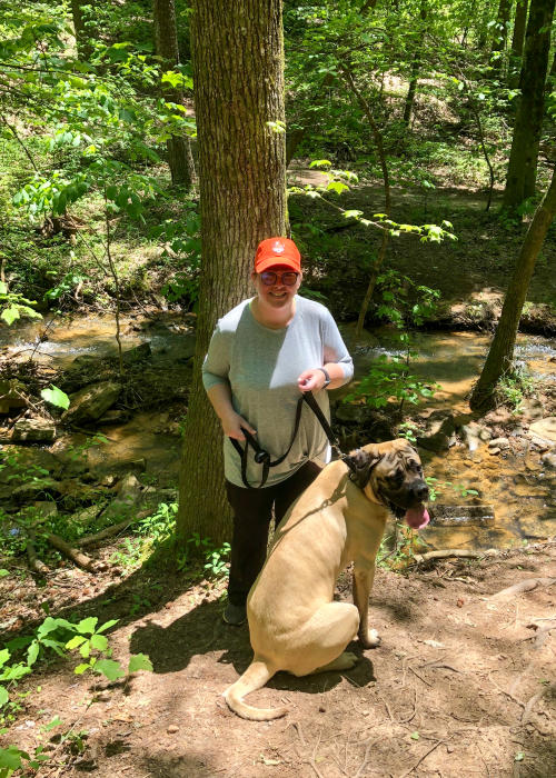A woman stands with a leashed dog beside a creek along Alum Hollow Trail, surrounded by lush green trees and foliage.