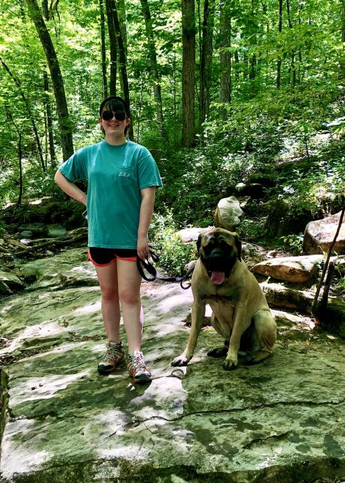 A woman stands with a leashed dog on a rocky section of Wildflower Trail, surrounded by green trees and foliage.