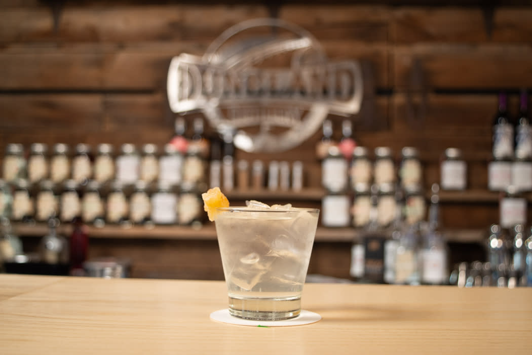 A clear cocktail with ice sits on a bar. In the background is a Duneland Distillery logo on the wall.