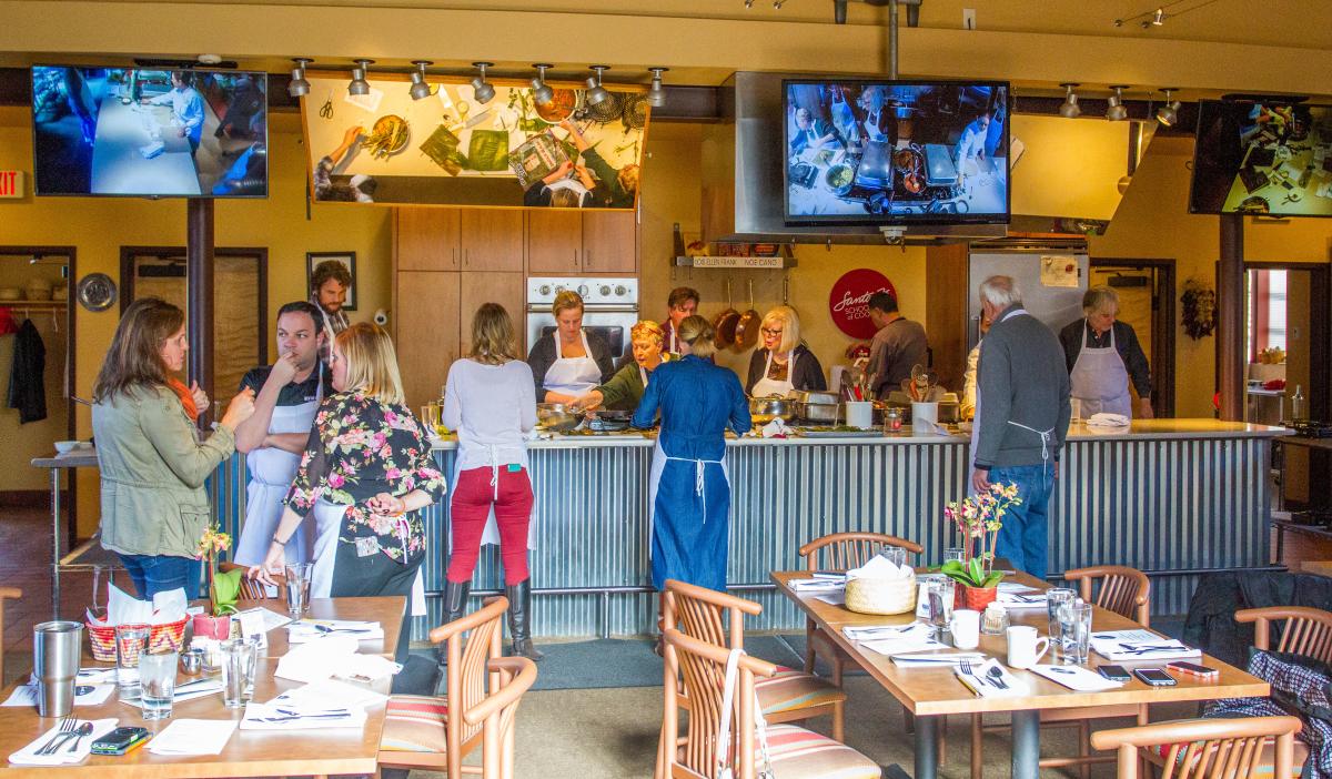A group of people standing in a kitchen space with tables and TV's hanging from the ceiling.