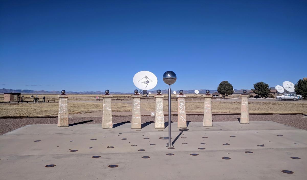 A metal sphere atop a slender post casts a shadow across a concrete surface dotted with markers.