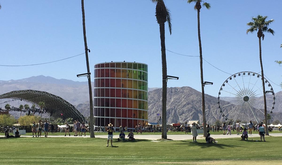 Spectra, a circular seven story structure with rainbow colored tinted windows stands in the middle of the Coachella Festival grounds by day with mountains in the background and a few festivalgoers scattered around.