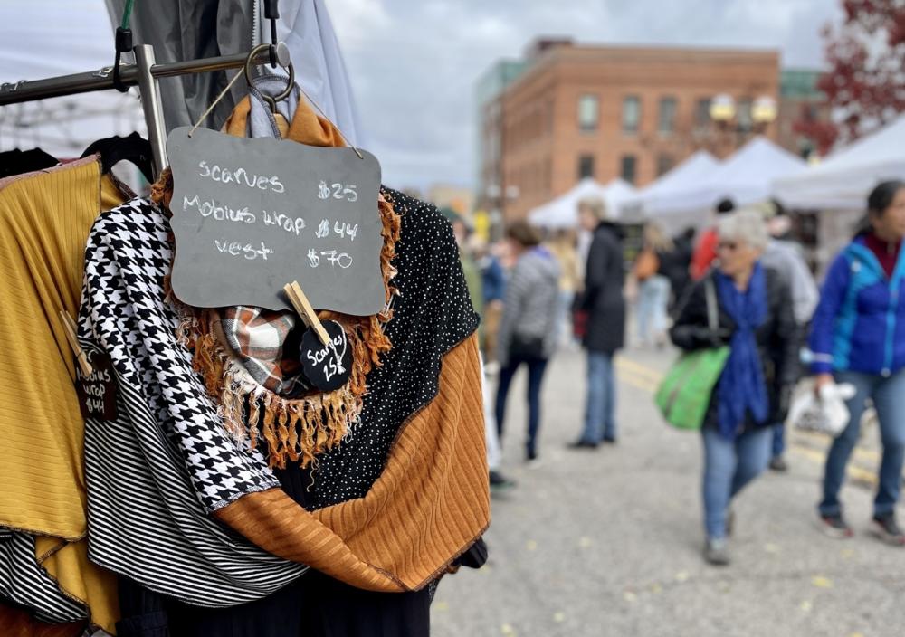 Close-up of scarves attached to hanger with prices listed in front of scarves. In the background, people walk past vendor booths filled with artwork lining the streets.