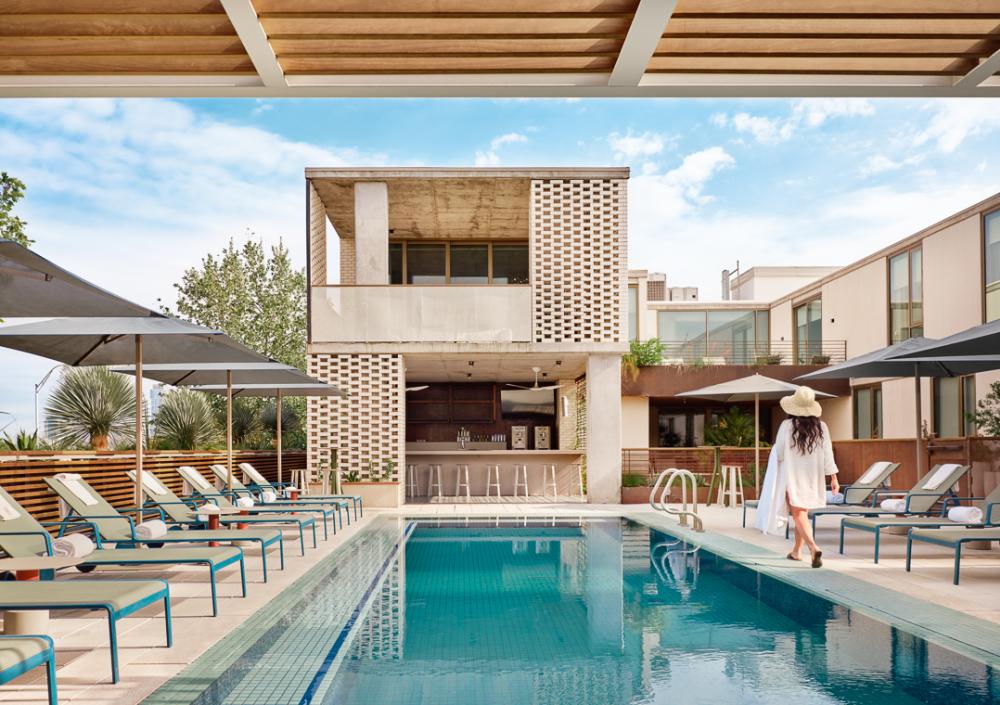 Woman in a shady sunhat and white swimsuit cover up walking alongside a rectangular pool lined with lounge chairs.