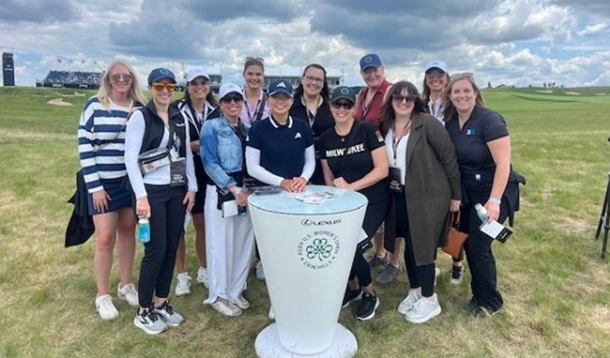 A group of thirteen people, including Visit Milwaukee team members and meeting planners, pose together outdoors at the 2025 U.S. Women’s Open at Erin Hills. They are standing around a branded Lexus event table on a grassy golf course under a partly cloudy sky.