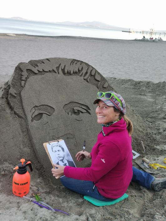 A woman sits on the beach, carving a face into the sand. She is wearing a magenta sweatshirt and jeans and looks over her shoulder at the camera.