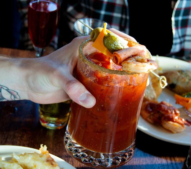 A hand holds a bloody mary that is topped with a small skewer with an olive, pickle, ham, and shrimp at Granite City