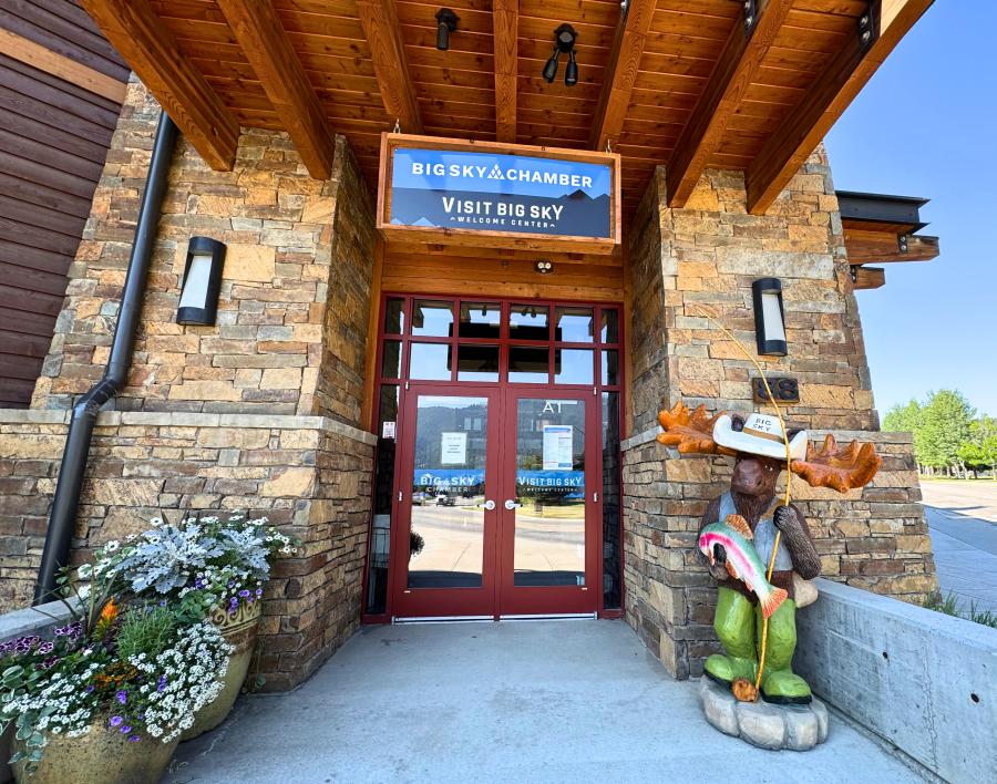 Big Sky Chamber/Visit Big Sky entrance with stone facade, red doors, floral planters, and a green moose statue with fishing gear.