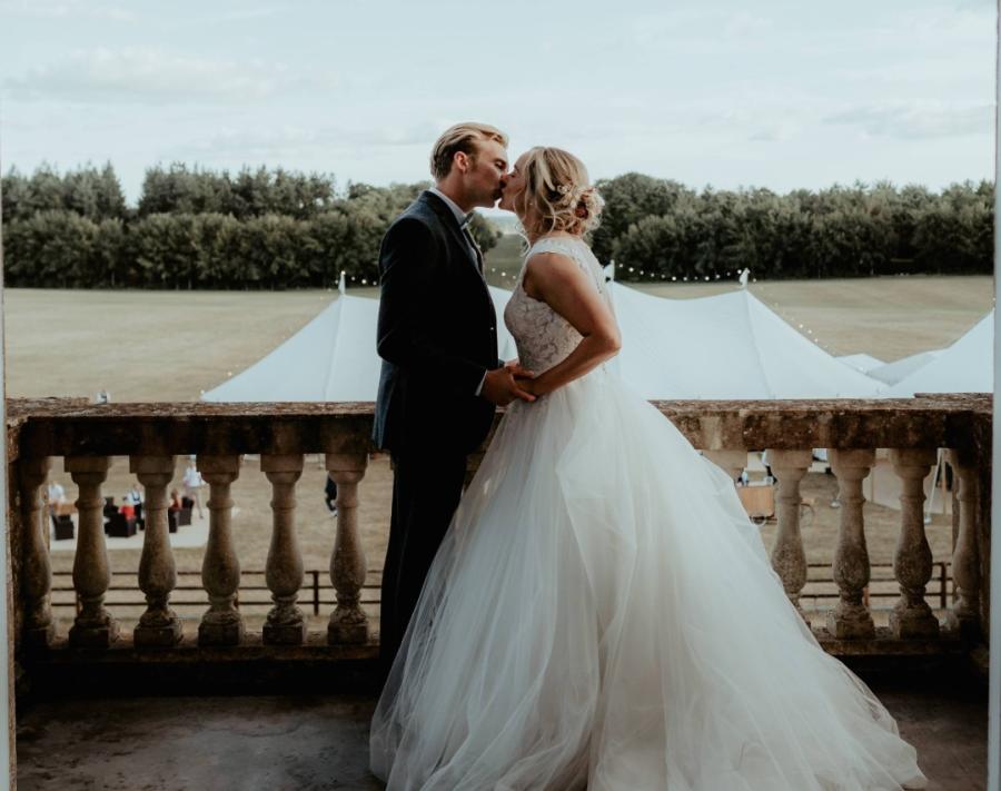 A bride and groom kiss on a balcony overlooking a wedding marquee and the grounds of the Badminton Estate