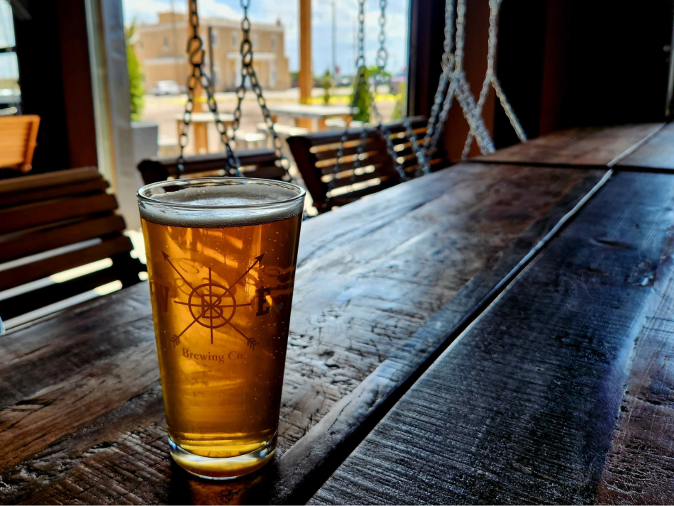 A pint of beer with the Westby Edge Brewing Company logo, placed on a rustic wooden table with swinging benches in the background, inviting a cozy atmosphere.