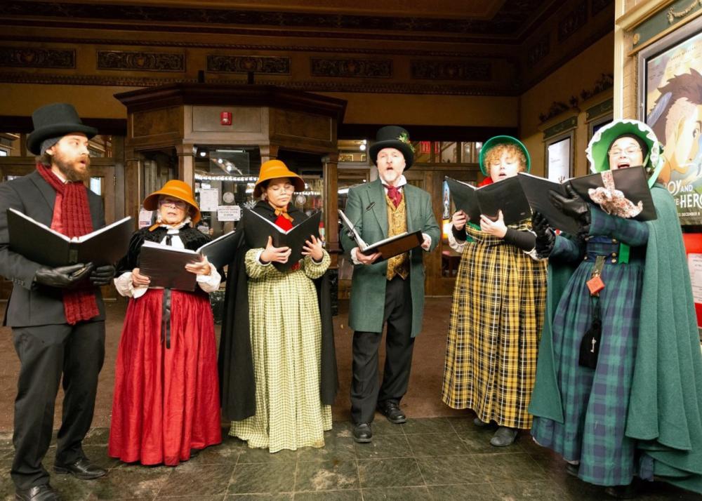 six individuals dressed up for holiday caroling, with music books open reading and singing together.