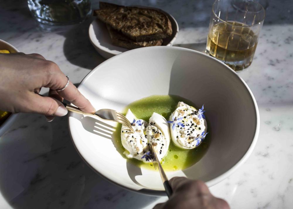 Moody image of hands holding knife and fork, slicing open a flower adorned ball of burrata cheese.
